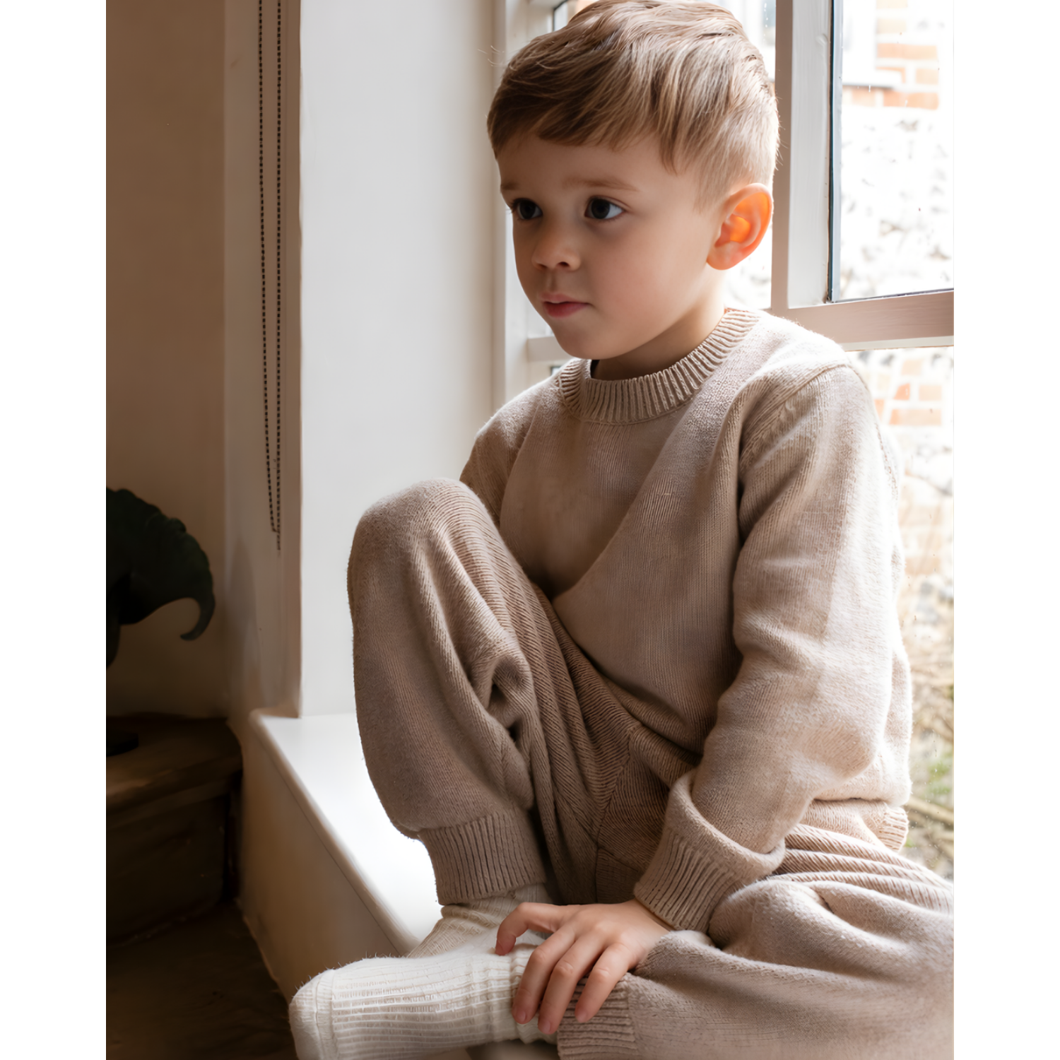 Child wearing a NELBEE beige sweater and pants sitting by a window
