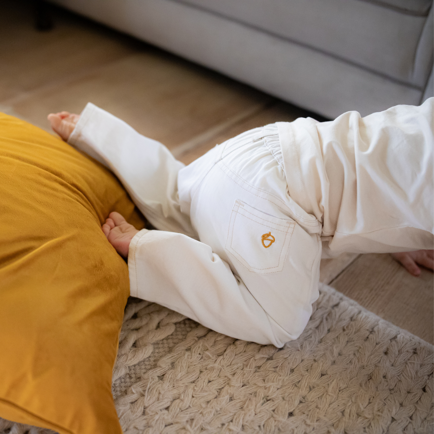 Child in NELBEE white pants crawling on a wooden floor next to a yellow pillow.