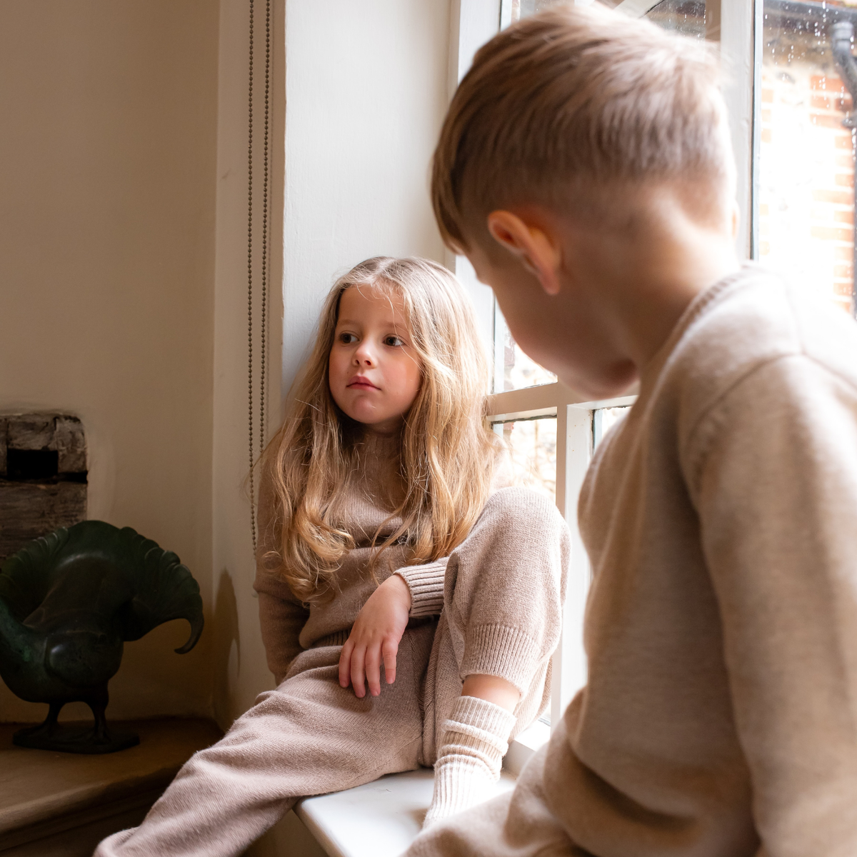 Two children sitting by a window, looking outwards wearing NELBEE Knitted sets.