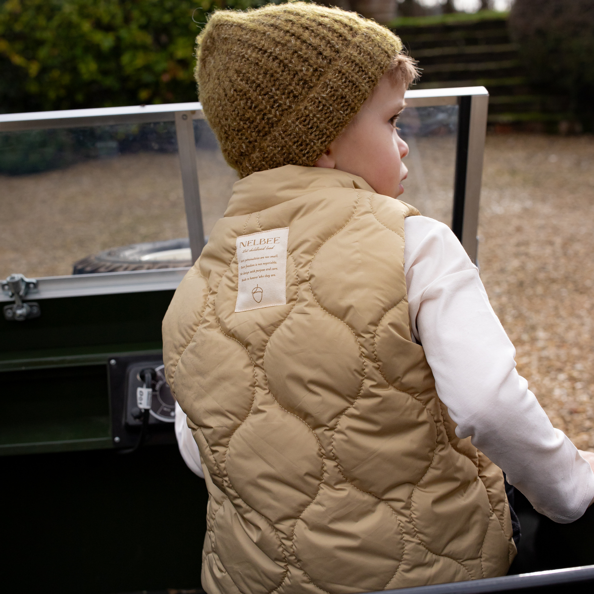 Child wearing NELBEE beige quilted vest and knitted hat sitting on a vehicle.