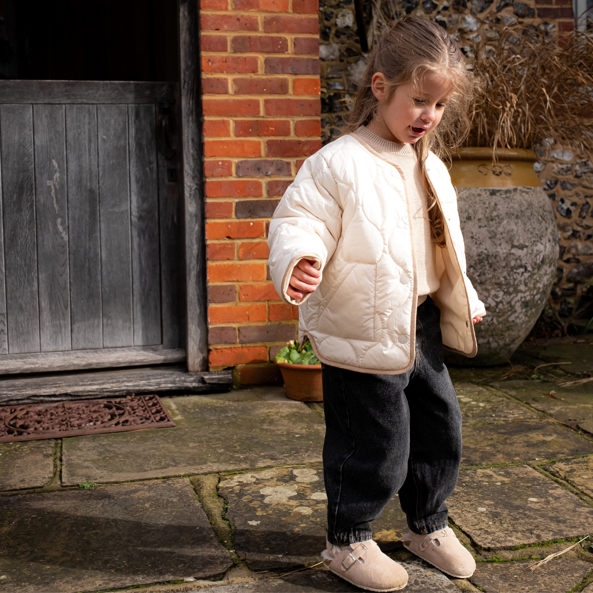 Girl in NELBEE beige quilted jacket standing on a stone path next to a brick building.