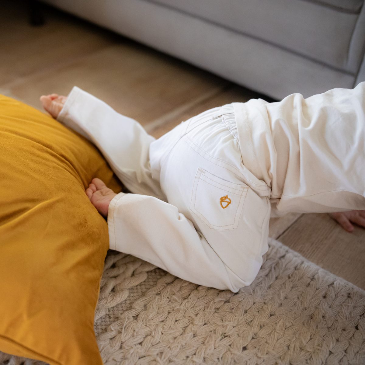 Child in NELBEE white pants crawling on a wooden floor next to a yellow pillow.